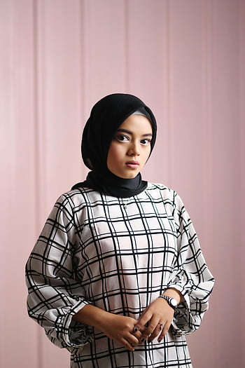 nainen, valkoinen ja musta ruudukon kuviomekko, musta hijab, seisoo lähellä seinää, tyylikäs muotokuva, Tyylikäs ruudullinen asu, sisällä, vaaleanpunainen tausta, muoti, vaatimaton tyyli, hijab-muoti