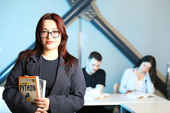 joven profesional, libro de python, escenario de clase, mujer sosteniendo un libro, Programación Python, estudiantes en segundo plano, ambiente educativo, atuendo profesional, ambiente de aprendizaje, lectura de libros, escena del aula