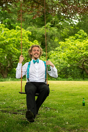 man in white dress shirt, black pants, sitting on swing, smiling young man, drink, lush park setting, bow tie, outdoor, green grass, trees, casual setting