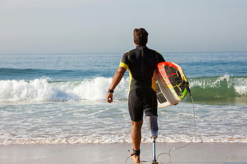 Amputeret surfer, Gå mod havet, surfbræt, ocean, Inspirerende billede, strand, bølger, våddragt, Protetisk ben, Adaptive sportsgrene, motiverende foto