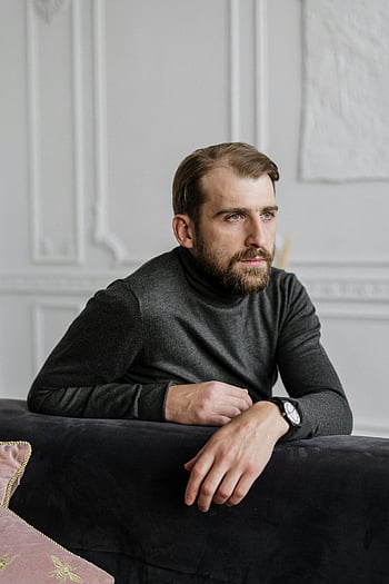 man in zwarte trui, Zittend op een zwarte bank, attente volwassen man, stijlvolle binnenomgeving, rustige sfeer, introspectieve sfeer, leunend op bank, binnenshuis portret, Stijlvolle man, zwarte bank, zwarte trui