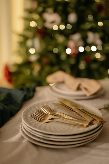 Christmas table setting, gold cutlery, white plates, festive table, Christmas tree backdrop, holiday decor, tableware, dining setup, elegant table, holiday table, stacked plates