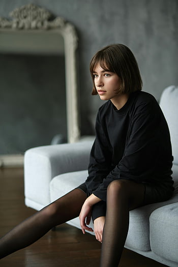 tonårsflicka, kontemplativ utseende, sitter på soffan, svart blus, inomhus porträtt, grå soffa, svarta tights, avslappnad pose, inomhusmiljö, ung kvinna, tankeväckande uttryck