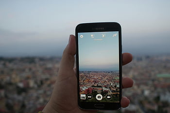 person, holding, black Samsung Galaxy, Android smartphone, taking picture, cityscape, blue sky, white sky, daytime, urban view, hand-held device