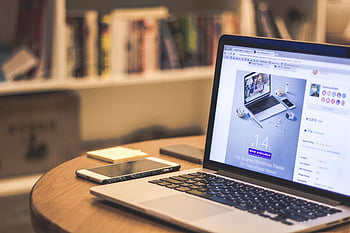 silver laptop, white smartphone, brown wooden table, modern workspace, cozy workspace, laptop displaying website, smartphone, books on shelves, workspace setup, tech workspace, home office setup