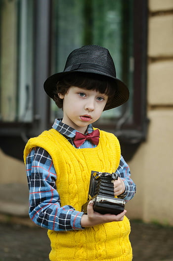 anak laki-laki memegang kamera, anak yang menawan, rompi kuning, kamera vintage, di luar rumah, topi hitam, kemeja kotak-kotak, dasi kupu-kupu merah, fotografer muda, gaya retro, fotografi anak