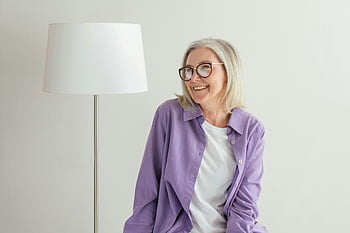 starsza kobieta, fioletowe długie rękawy, uśmiechnięty, okulary, wnętrz, biała lampa, radosny, fioletowa koszulka, posiedzenie, wewnątrz, białe tło