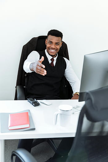 man in black vest, sitting behind desk, smiling professional, office setting, welcoming handshake, business attire, office environment, professional man, desk setup, office desk, handshake gesture