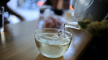 clear glass pitcher, pouring liquid, clear glass cup, steaming herbal tea, transparent glass, wooden table, close-up shot, tea pouring, glass teapot, hot beverage, tea time