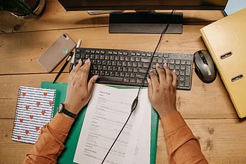 person using computer, keyboard, tidy workspace, wooden desk, office supplies, papers, computer mouse, workspace organization, desk setup, hands typing, office environment