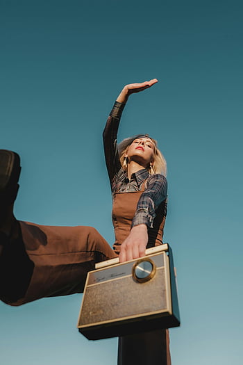 Froschperspektive, Frau im braunen Jumper, Retro -Radio halten, Vintage -Kleidung, klarer blauer Himmel, kreatives Posen, stilvolle Frau, Retro -Radio, Vintage-Stil, Foto im Freien, blauer himmel hintergrund
