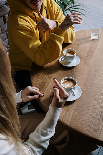 man and woman, communicating, hand sign, sign language, coffee, intimate setting, indoor, table, conversation, casual, yellow hoodie