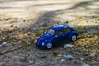 blue beetle car, miniature toy car, Volkswagen Beetle, sunlit gravel path, nature surroundings, outdoor scene, blue car, toy vehicle, gravel path, natural setting, miniature model