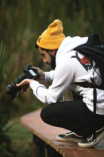 mies, jolla kamera, musta dslr-kamera, rento valokuvaaja, rypistyminen puisella polulla, puistovalokuvaus, kameran vaihde, Ulkokuvaus, keltainen pipo, valkoinen huppari, mustat housut, lenkkarit
