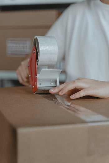 tape dispenser, cardboard box, sealing box, indoor setting, packing supplies, person packing, tape application, box sealing, packaging process, hand holding tape, cardboard packaging