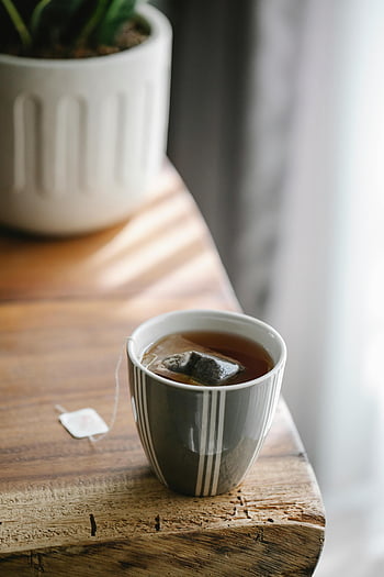 hot tea, grey ceramic cup, striped cup, teabag, rustic wooden table, warm morning, tea time, cozy drink, indoor setting, natural light, potted plant
