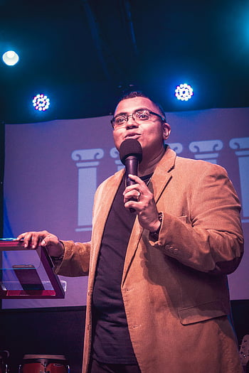 man in brown coat, holding microphone, speaking into microphone, stage performance, under spotlights, public speaking, event speaker, formal attire, microphone speech, stage event, spotlight presentation
