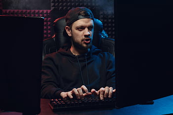 man with cap, typing on keyboard, dimly lit room, concentrating on screen, man wearing cap, hoodie, computer screen, gaming chair, indoor setting, focused expression, keyboard typing