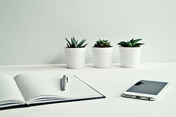 white ceramic pots, green leaf plants, open notebook, click pen, minimalist workspace, smartphone, potted succulents, clean desk, white table, office setup, productivity space