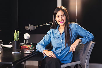 woman, denim top, podcast studio, smiling woman, sitting at desk, microphones, studio setting, professional photo, casual attire, office environment, desk setup