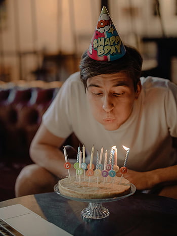 birthday celebration, man blowing candles, home birthday party, birthday cake, candles on cake, happy birthday hat, indoor celebration, birthday party, birthday cake decoration, man celebrating birthday, home party