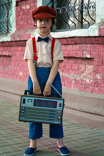 młody chłopiec, vintage radio, moda retro, ceglana ściana, scena na zewnątrz, czerwony dom, muszka, szelki, czapka z daszkiem, spodenki jeansowe, niebieskie buty