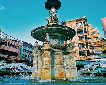 betonová fontána, sochařská fontána, městské budovy, jasné modré nebe, Urban Fountain, venkovní socha, veřejné umění, vodní prvek, architektonické mezník, městské krajiny, panoráma města