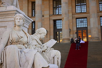 dvě ženy sochy, elegantní mramorové sochy, historické budovy, červený koberec, klasická scéna, mramorová socha, architektonické detaily, venkovní umění, kulturní památka, kamenné postavy, červený koberec událost