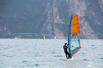 windsurf, homem de roupa de mergulho preta, veleiro laranja e azul, lago cênico, entusiastas da aventura, dia, Experiência emocionante, Esportes Aquáticos, atividades ao ar livre, barco a vela, windsurfista