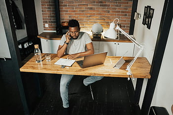 Mann arbeitet von zu Hause aus, Innenministerium, Heimarbeit, junger Mann, Laptop, Telefon, Schreibtisch, Lebensstil arbeiten, Heimarbeitsplatz, Ziegelwand, Holztisch