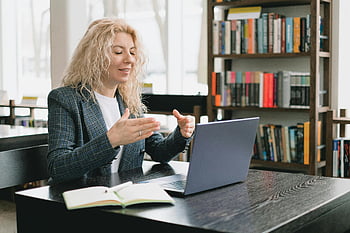 Video-Chat, Laptop, Bibliothek, blonde Frau, professionelles Engagement, Bildungsumgebung, lächelnde Frau, Bücherregal, Online-Meeting, Studienumfeld, Heimarbeit