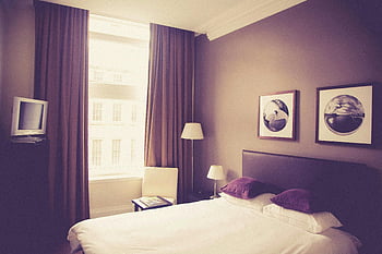 white bed linen, white table lamp, vintage style bedroom, natural light, cozy decor, purple curtains, framed wall art, bedside table, floor lamp, window view, hotel room