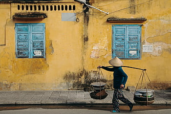 street vendor, traditional attire, aged yellow building, blue windows, merchant walking, street scene, Vietnamese culture, carrying goods, urban life, Southeast Asia, daily life
