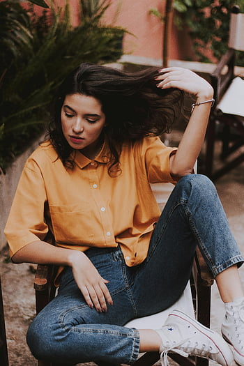 woman sitting, wooden chair, holding hair, stylish woman, casual attire, outdoors, relaxed vibes, fashionable vibes, yellow shirt, blue jeans, white sneakers