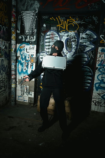 black jacket, mask, silver briefcase, gun, graffiti-covered wall, person in disguise, urban setting, dark alley, street art, mysterious figure, crime scene