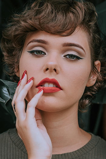 woman touching face, right hand, bold red lipstick, elegant makeup, classic beauty, close-up portrait, red nails, vintage style, soft lighting, natural skin, expressive eyes