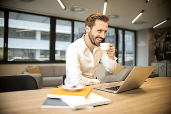 depth of field, man sitting, holding cup, modern office, coffee break, laptop, books, table, chair, professional setting, work environment