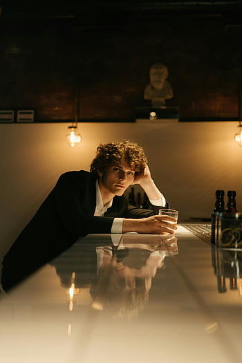 thoughtful man, black suit, stylish bar, holding drink, warm lighting, bar setting, reflective surface, casual pose, indoor scene, modern decor, ambient light