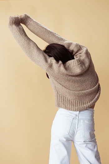 woman stretching, brown sweater, white denim jeans, minimalist background, cozy sweater, arms overhead, beige background, fashion photography, casual style, women's fashion, lifestyle image