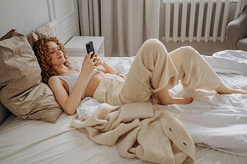 Frau im weißen Erntepunkt, auf dem Bett liegen, mit Smartphone, Gemütliche Morgenszene, Entspannen im Bett, Smartphone, Bett, Weißes Erntepunkt, Gemütliche Szene, entspannend, Smartphone -Nutzung