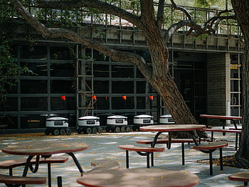 meja kafetaria yang tidak terpakai, robot pengiriman otonom, halaman modern, halaman kosong, Tabel luar ruangan, pohon, meja piknik, robot pengiriman, bangunan modern, tempat duduk di luar ruangan, meja kosong