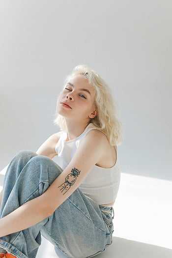 woman in white top, blue denim jeans, blonde woman, sitting, winking, tattoo, arm tattoo, casual style, portrait, white background, casual fashion