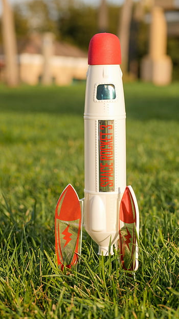 toy rocket, close-up shot, green grass, daylight, blurred background, space rocket, outdoor scene, toy spaceship, red and white, toy model, toy design