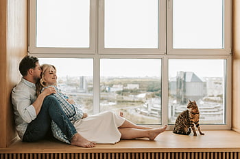 pregnant couple, relaxing by window, affectionate couple, cozy moment, cat nearby, window view, pregnant woman, couple sitting, indoor scene, pet cat, cityscape background