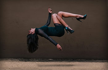 woman in levitation, long-sleeved mini dress, elegant dress, textured wall, levitation pose, high heels, black dress, artistic photography, dynamic pose, female model, urban backdrop