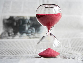 hourglass, clear glass, red sand, pink sand, flowing sand, close-up, newspaper background, transparent hourglass, sand timer, time measurement, decorative hourglass
