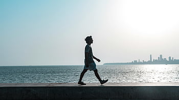 man walking, body of water, Mumbai coastline, sunset, cityscape backdrop, silhouette, seaside walk, urban skyline, waterfront scene, evening stroll, coastal path