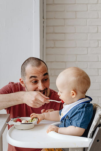 man feeding baby, red shirt, blue shirt, high chair, brick wall, indoor, father, baby, feeding time, family moment, love