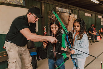 archery, young girls, male instructor, indoor training, learning, concentration, diverse children, cute, attentive, bow and arrow, trainer