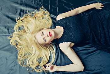 Frau, die auf dem Bett liegt, elegantes Modeporträt, blonde Frau, luxuriöses schwarzes Kleid, anmutig liegen, Modefotografie, blondes Haar, roter Lippenstift, Bettporträt, High Fashion, eleganter Stil
